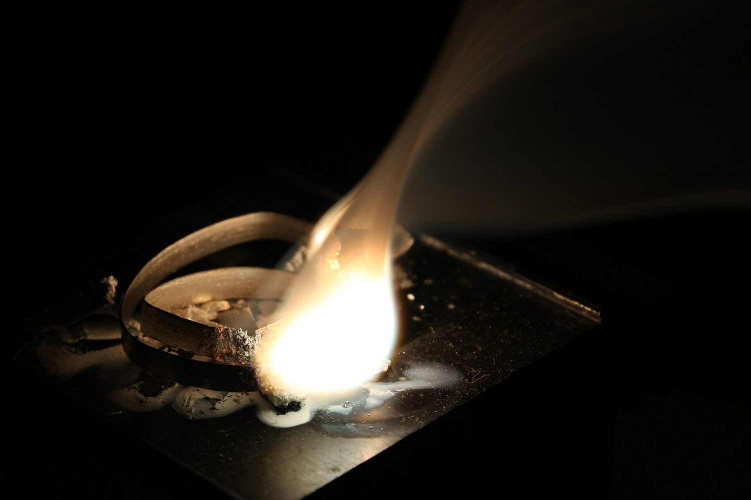 A strip of magnesium held in pliers burns with a dazzling white light, giving off white smoke of magnesium oxide