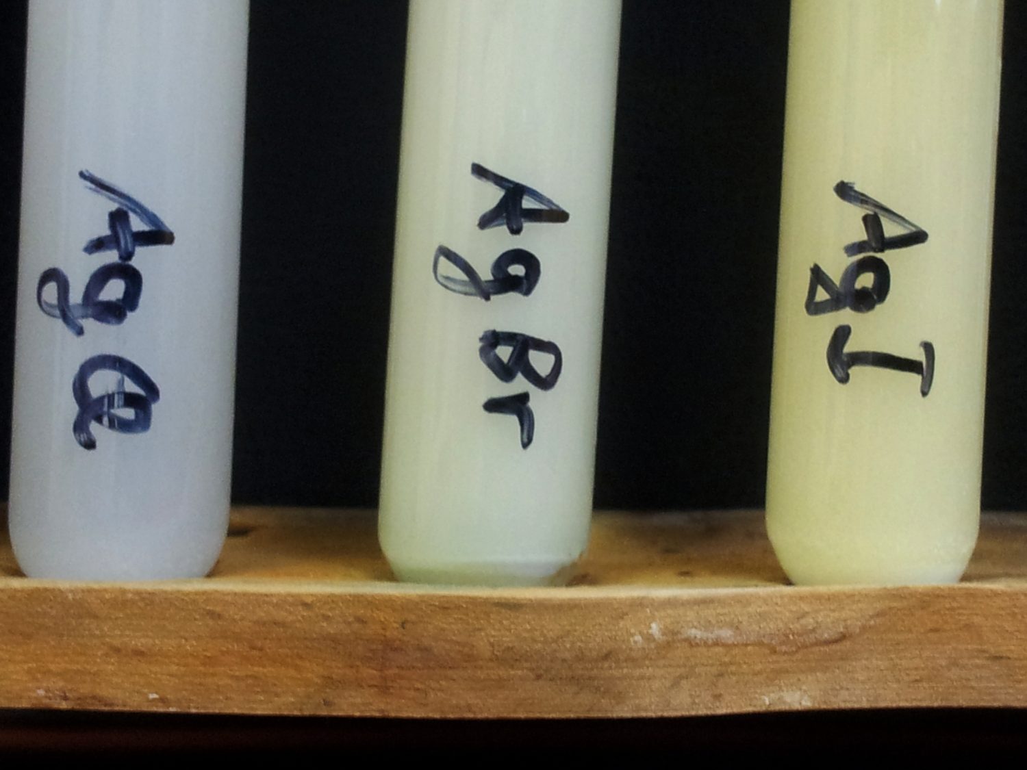 Three real test tubes in a wooden rack, labelled AgCl, AgBr and AgI, holding a white, a pale cream and a pale yellow precipitate from left to right
