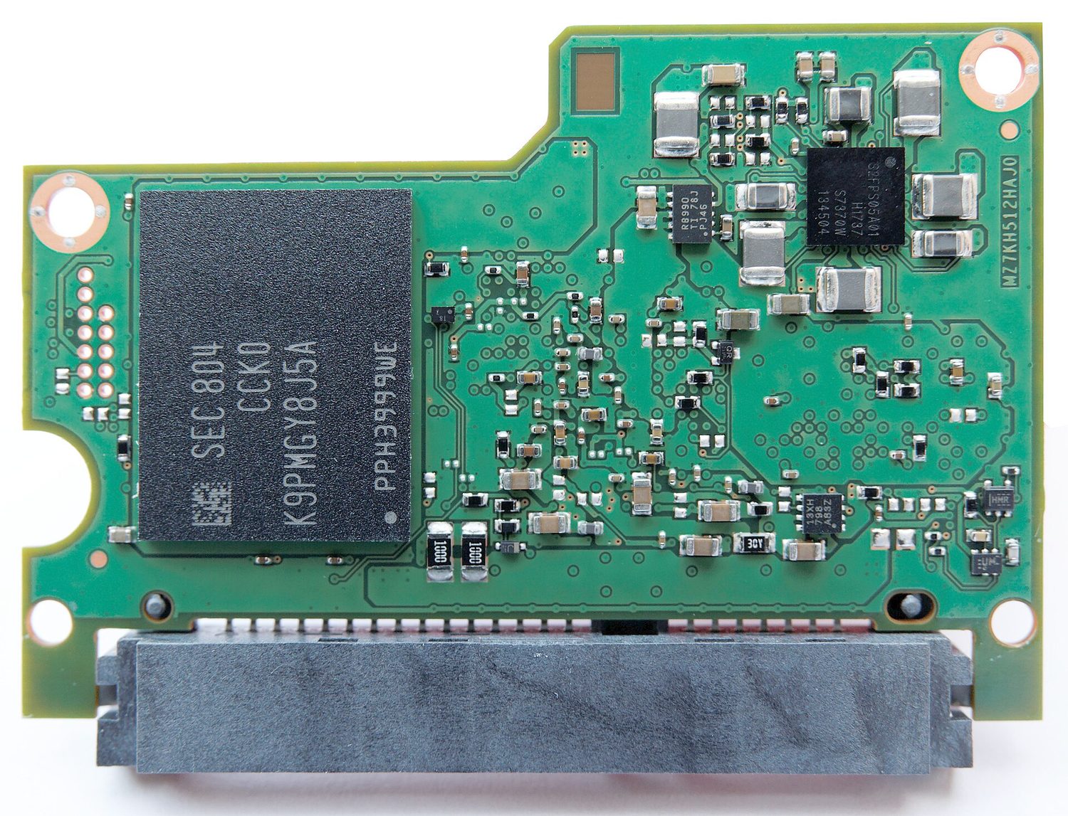 The opened circuit board of a solid-state drive on a white background: a large black flash-memory chip on the left, a smaller controller chip, many tiny components, and a flat SATA connector along the bottom edge — no platters or moving parts