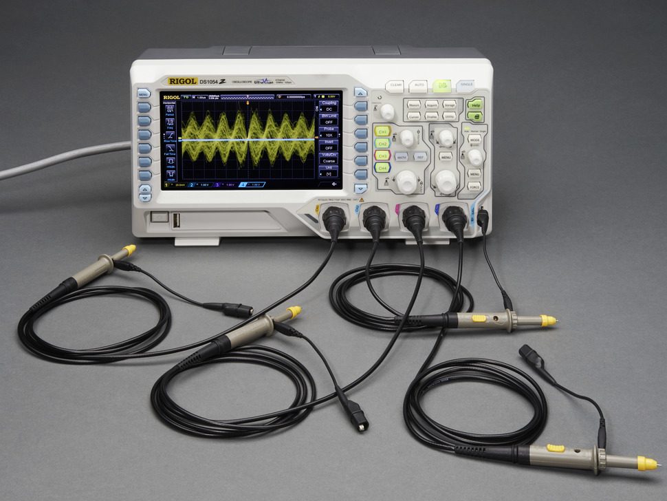 A modern digital oscilloscope with a grid screen showing a yellow voltage signal against time, a row of control knobs and buttons, and four probe leads plugged into the input sockets