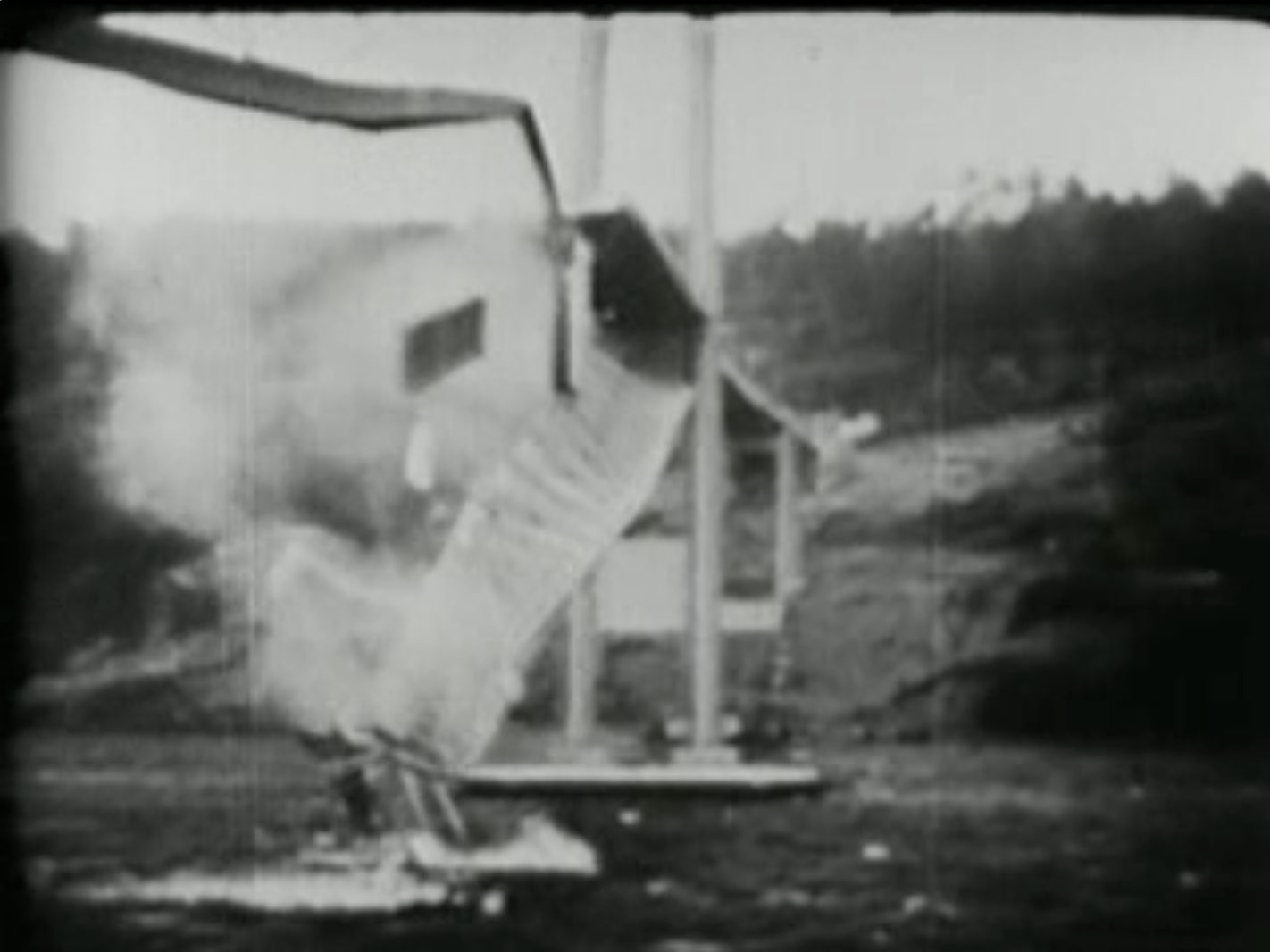 A black-and-white photograph of the Tacoma Narrows Bridge roadway twisted far over to one side while a man clings to it