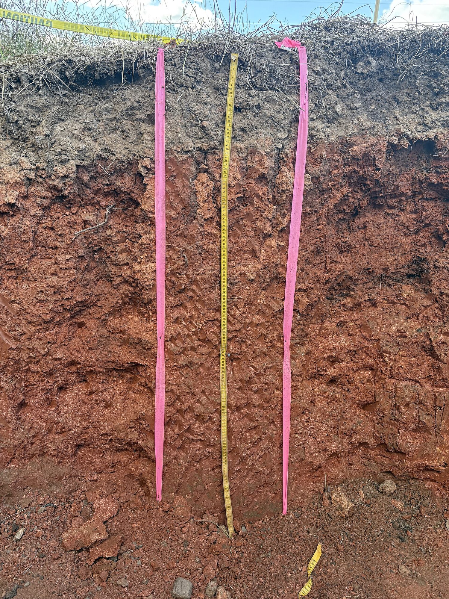 A vertical pit cut through soil, showing a dark topsoil band over reddish-brown mineral layers, with a tape measure for scale