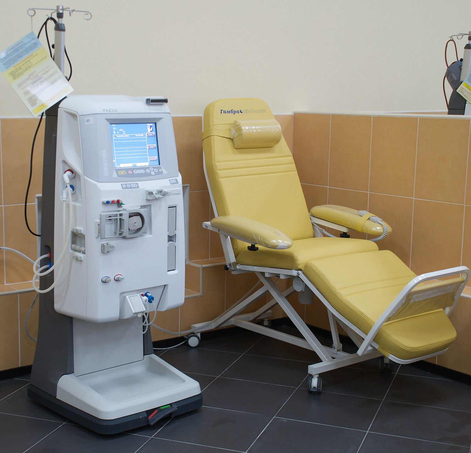 A modern dialysis machine standing next to a patient's chair in a clinic; tubes carry the patient's blood through the machine, which removes waste and excess water and returns the clean blood