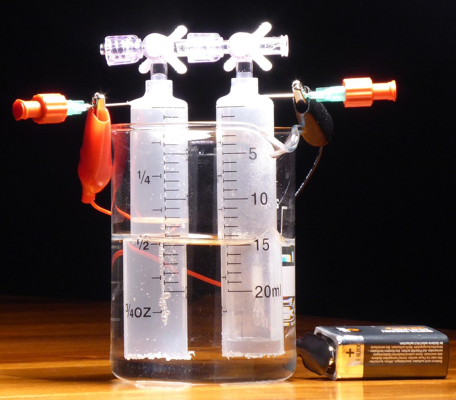 A small electrolysis demonstration: two upturned graduated tubes stand in a beaker of water, wired to a 9-volt battery, with gas collected at the top of each tube and about twice as much gas in one tube as the other