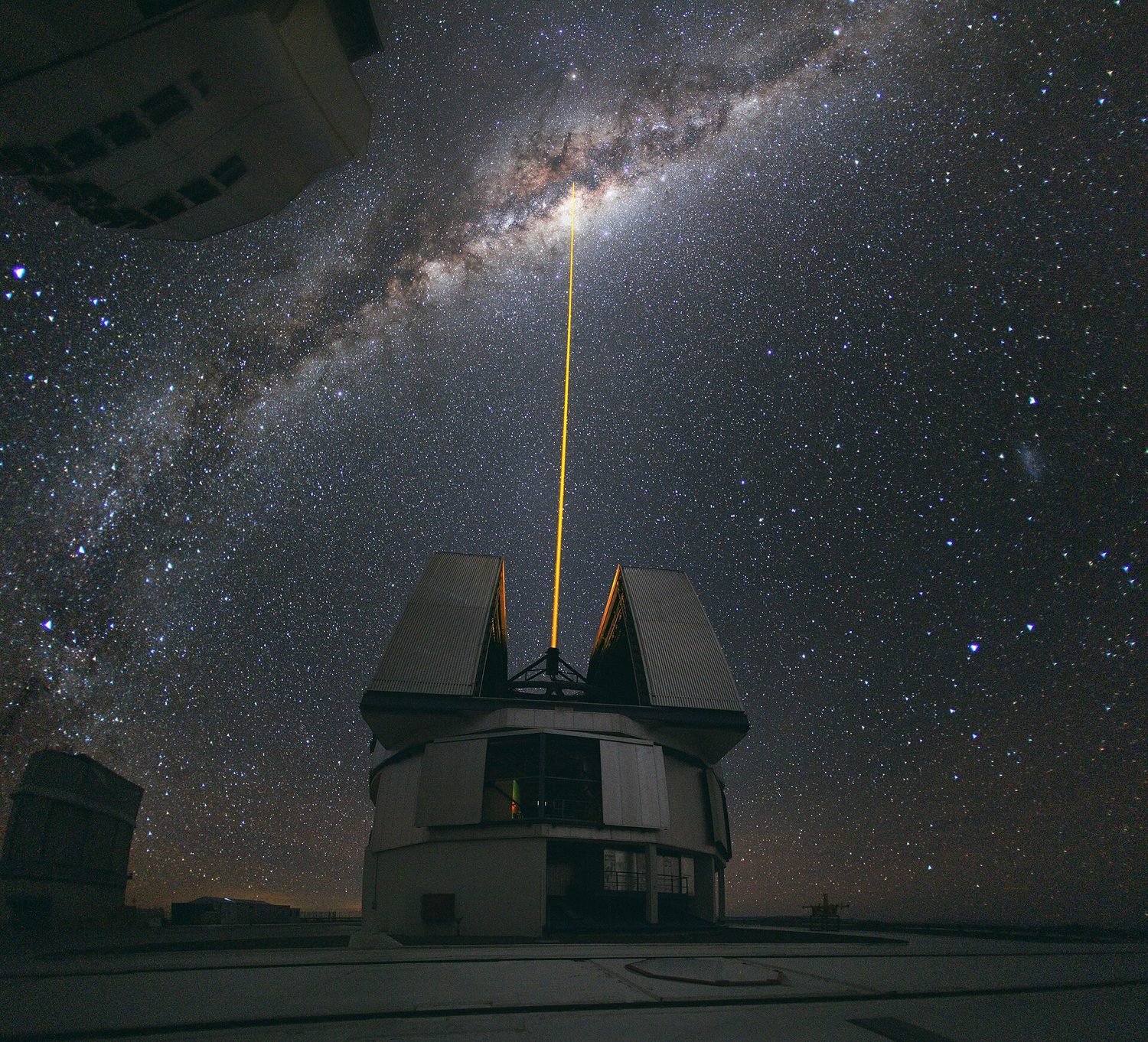 A large telescope on a mountain at night fires a thin yellow laser straight up towards the glowing band of the Milky Way